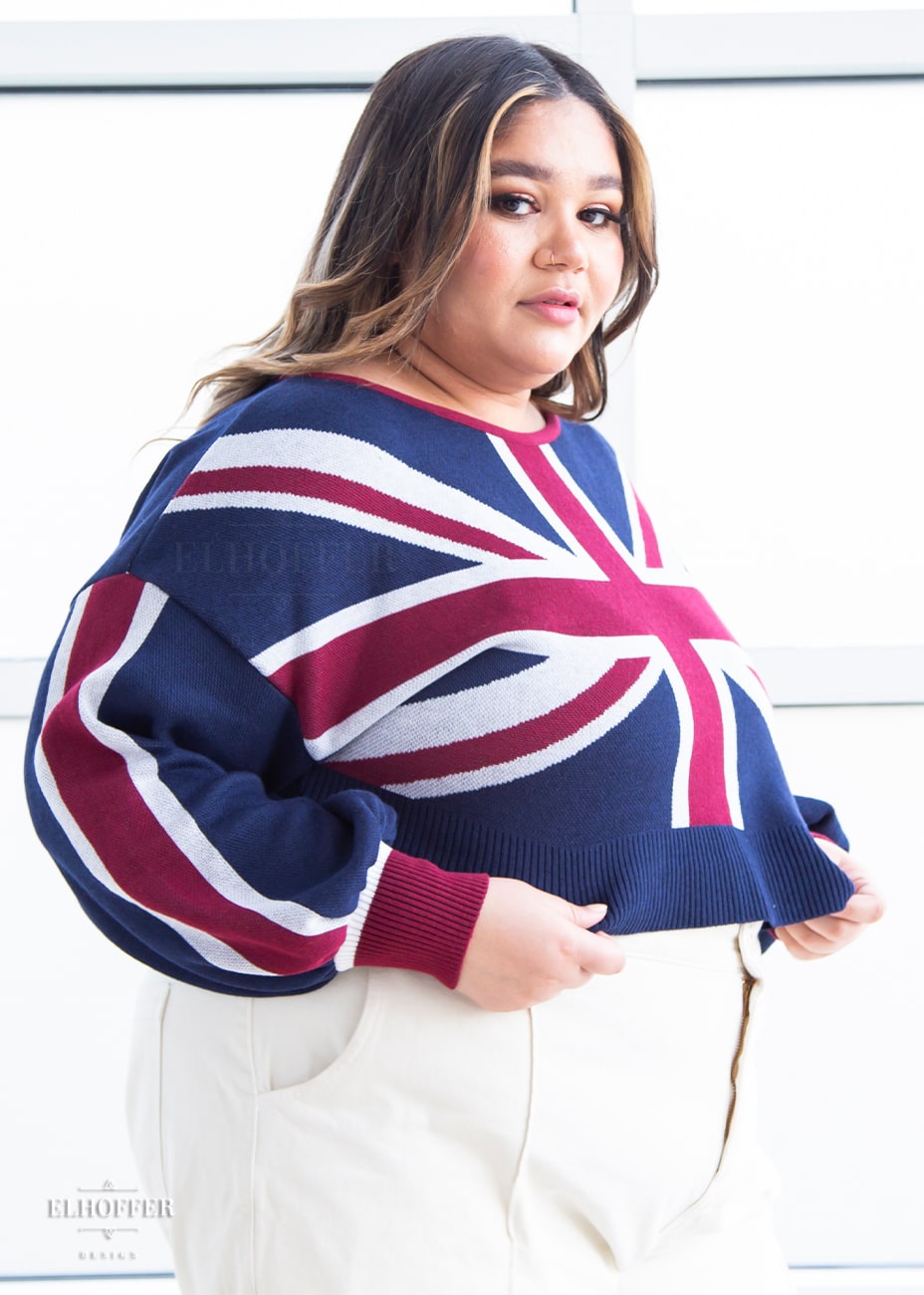 Cori, a sun kissed skinned 2xl model with long balayage hair, is smiling while wearing a cropped oversize sweater with a Union Jack design and long billowing sleeves.