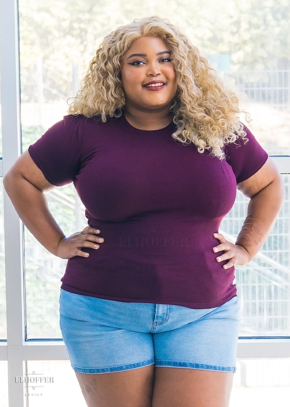 Jade, a medium dark skinned 2XL model with curly blonde hair, is wearing a dark cherry knit short sleeve tee style top. The top has a high round neckline and the hem of the top falls to the hips. The shoulders are slightly gathered.