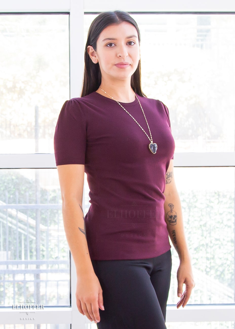 Alexandra, a light olive skinned S model with long dark brown hair, is wearing a short sleeve light weight dark reddish purple knit top. The top hits about mid hip in length and the sleeves have pleated gathering at the shoulders.