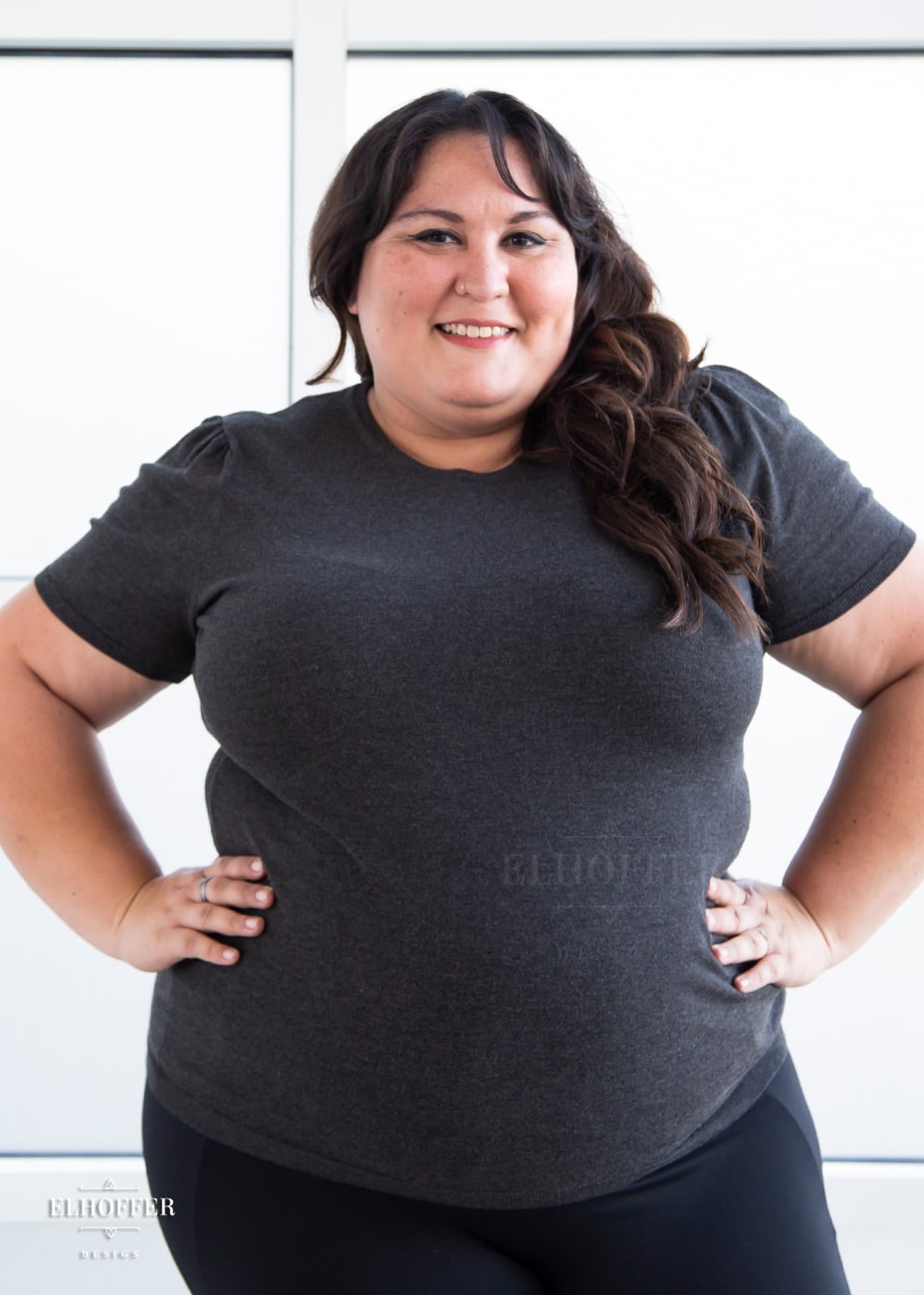 Alysia, a sun kissed skin 2xl model with long wavy dark brown hair, is smiling while wearing a short sleeve light weight charcoal grey knit top. The top hits about mid hip in length and the sleeves have pleated gathering at the shoulders.