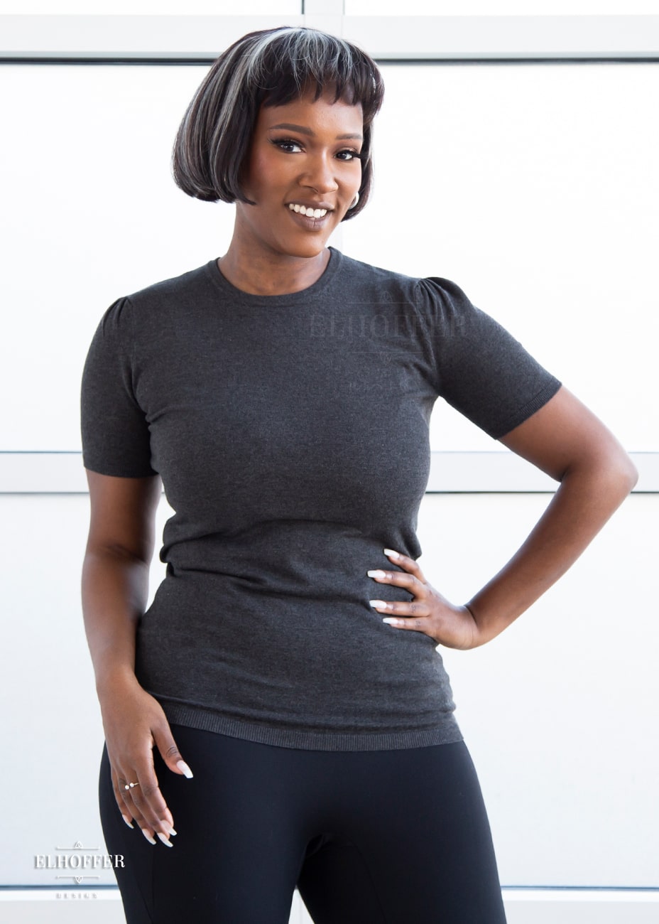 Lynsi, a medium dark skinned model with short black and white hair, is smiling while wearing a short sleeve light weight charcoal grey knit top. The top hits about mid hip in length and the sleeves have pleated gathering at the shoulders.