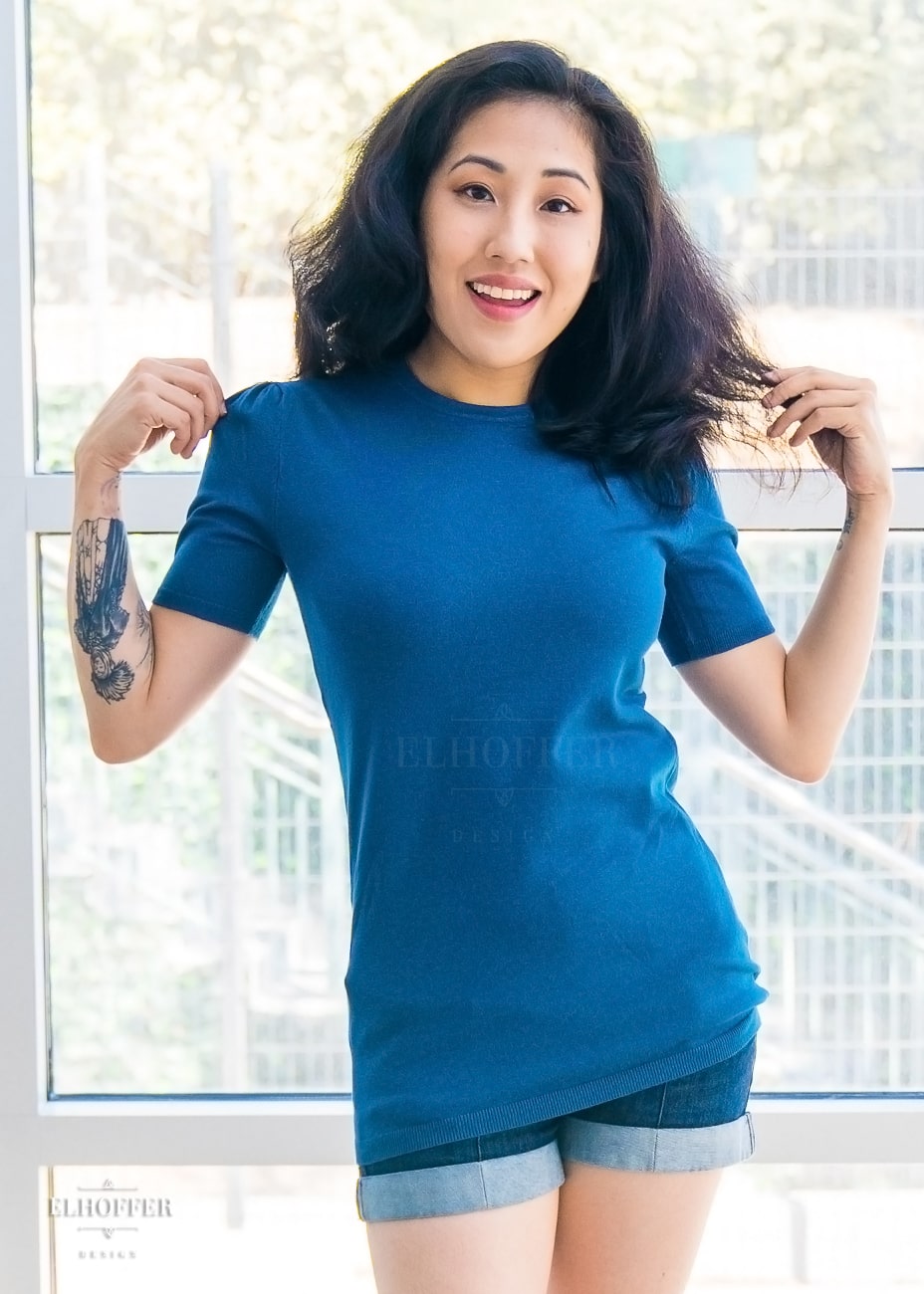 Kate, an olive skinned XS model with long dark hair, is wearing a blue knit short sleeve tee style top. The top has a high round neckline and the hem of the top falls to the hips. The shoulders are slightly gathered.