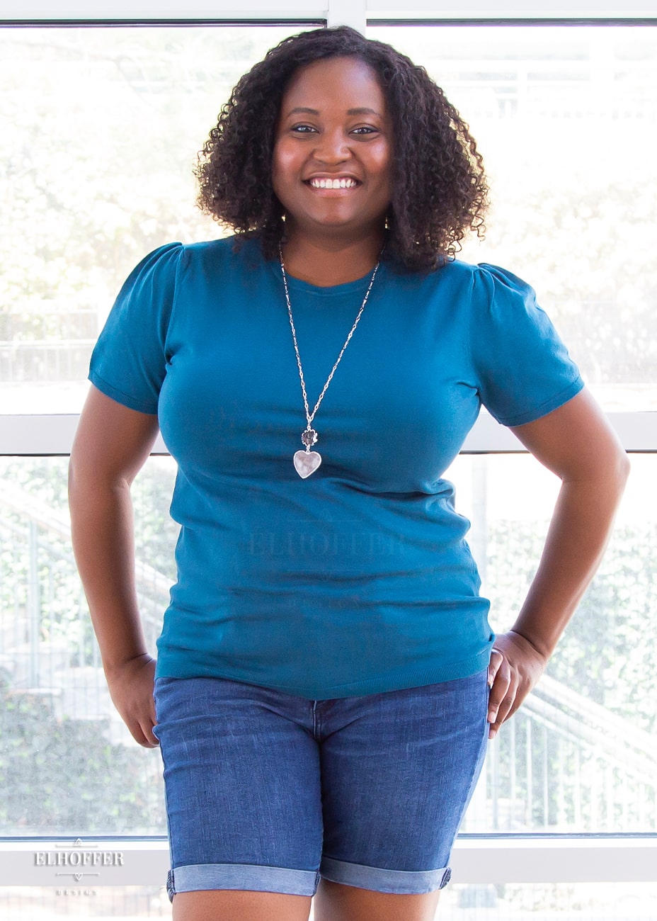 Maydelle, a medium dark skinned XL model with dark shoulder length tight curly hair, is smiling while wearing a short sleeve light weight peacock teal knit top. The top hits about mid hip in length and the sleeves have pleated gathering at the shoulders.