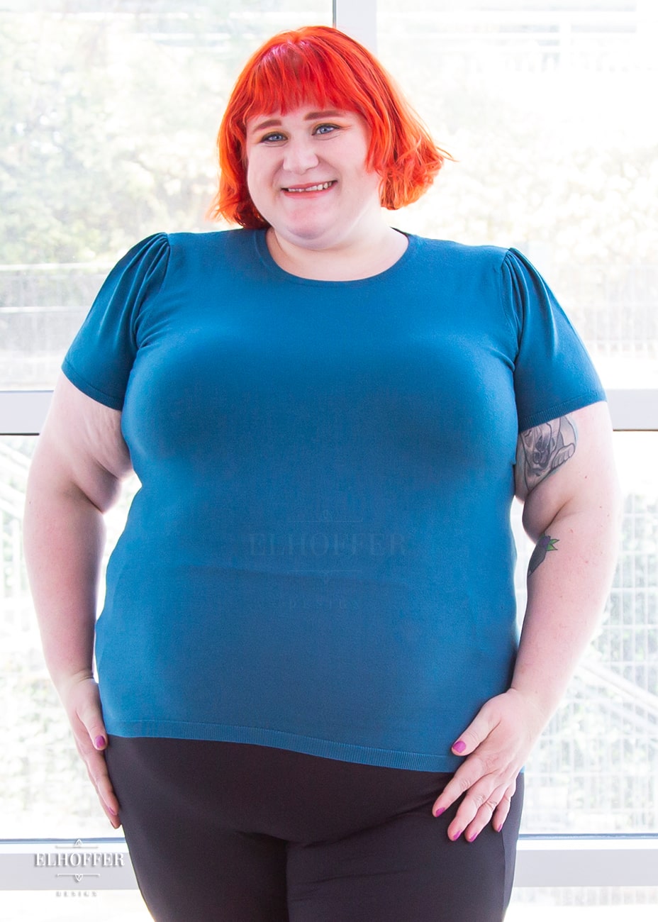 Logan, a fair skinned 3xl model with short orange hair with bangs, is smiling while wearing a short sleeve light weight peacock teal knit top. The top hits about mid hip in length and the sleeves have pleated gathering at the shoulders.