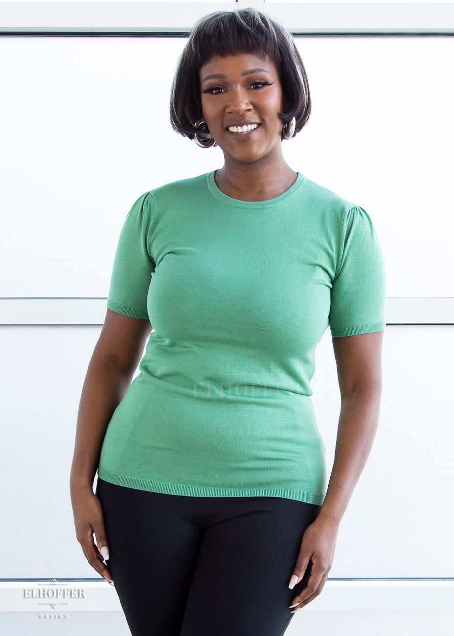 Lynsi, a medium dark skinned model with short black and white hair, is smiling while wearing a short sleeve light weight sage green knit top. The top hits about mid hip in length and the sleeves have pleated gathering at the shoulders.