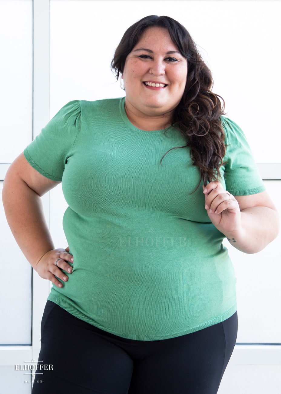 Alysia, a sun kissed skin 2xl model with long wavy dark brown hair, is smiling while wearing a short sleeve light weight sage green knit top. The top hits about mid hip in length and the sleeves have pleated gathering at the shoulders.