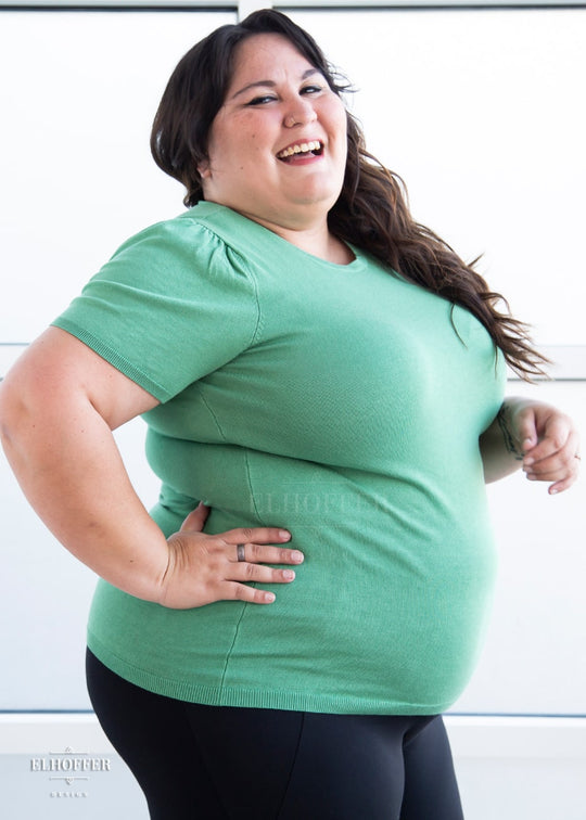 A side view of Alysia, a sun kissed skin 2xl model with long wavy dark brown hair, caught mid laugh while wearing a short sleeve light weight sage green knit top. The top hits about mid hip in length and the sleeves have pleated gathering at the shoulders.