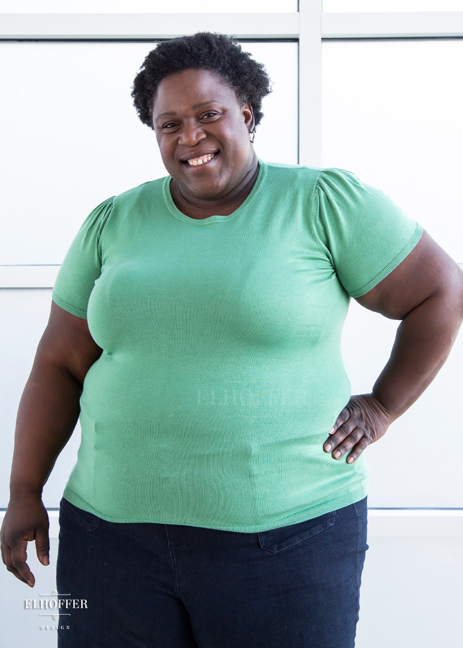 Adalgiza, a medium dark skinned 4xl model with short dark super curly hair, is smiling while wearing a short sleeve light weight sage green knit top. The top hits about mid hip in length and the sleeves have pleated gathering at the shoulders.