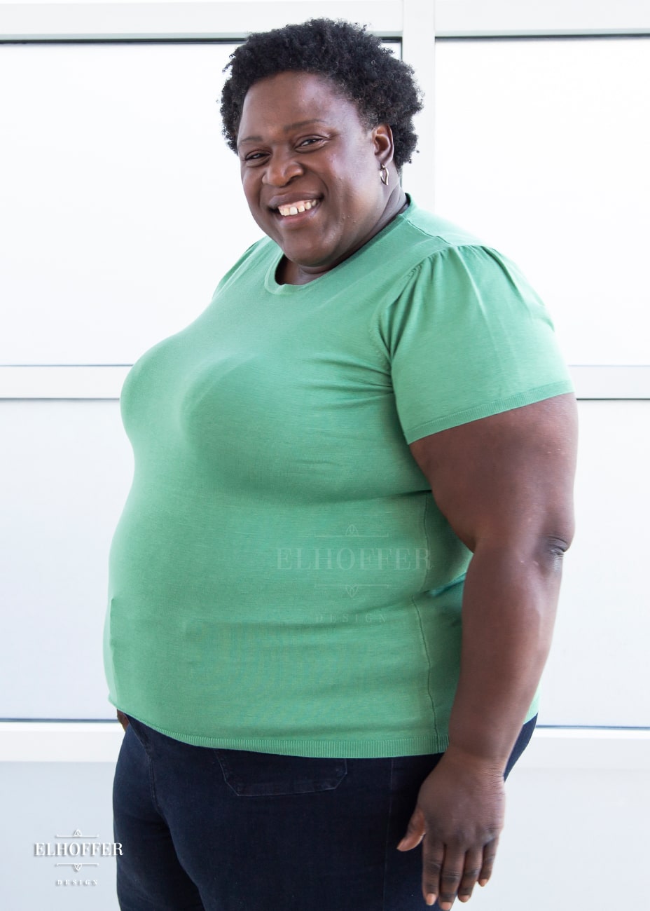 Adalgiza, a medium dark skinned 4xl model with short dark super curly hair, is smiling while wearing a short sleeve light weight sage green knit top. The top hits about mid hip in length and the sleeves have pleated gathering at the shoulders.