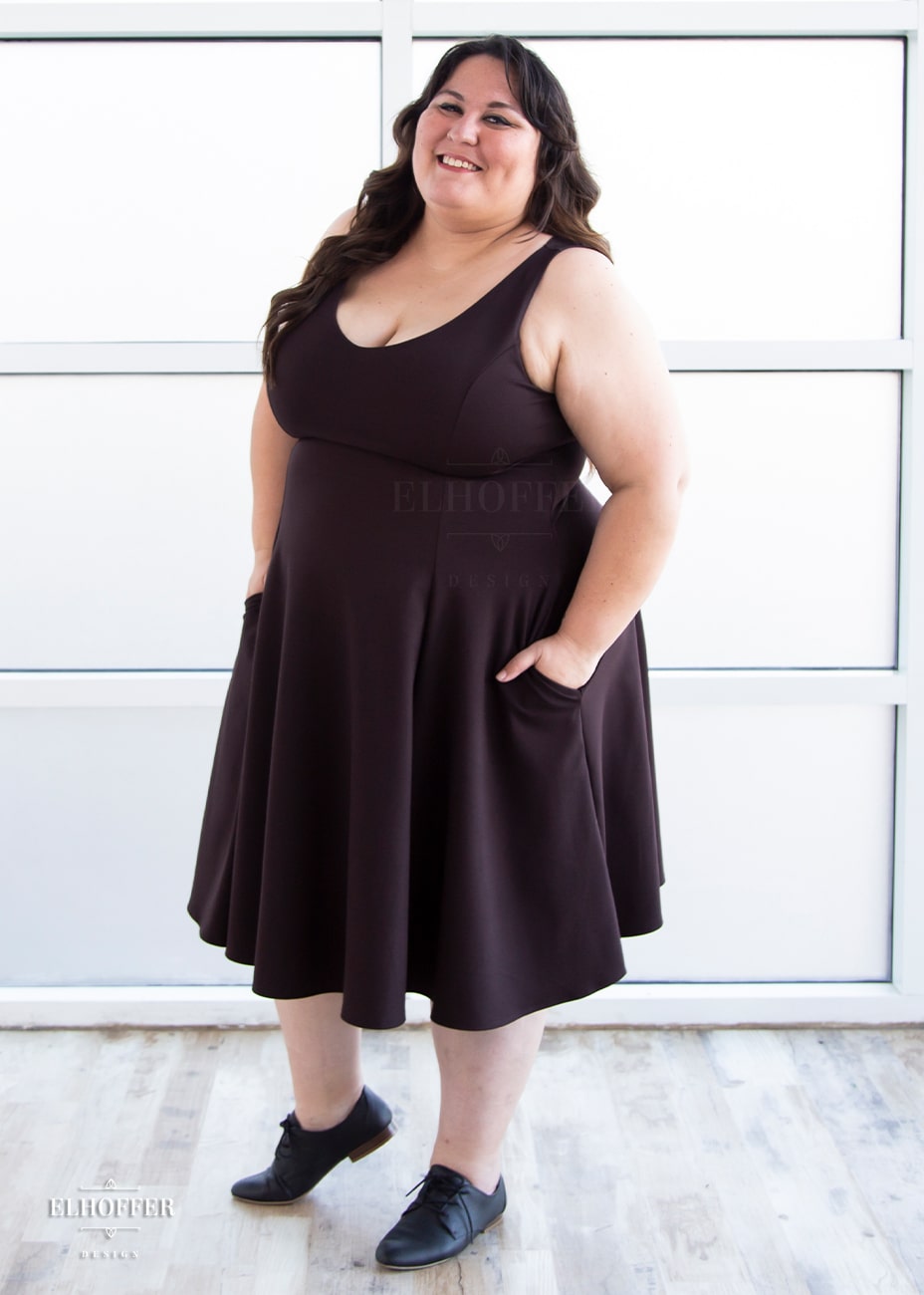 Alysia, a sun kissed skin XL model with long wavy dark brown hair, is smiling while wearing a reversible knee length chocolate brown dress with pockets.  The dress has a v neck on one side and a boat neck on the other and princess seams.