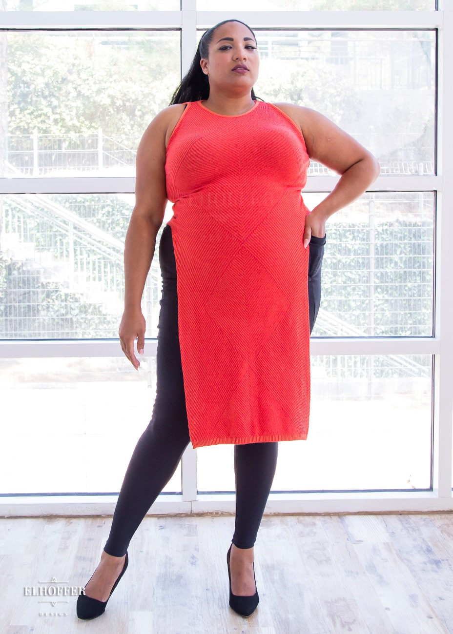 Tas, a medium dark skinned size 2XL model with long dark hair, is wearing a pullover cropped sleeveless knit top with front and back flaps in a medium orange.