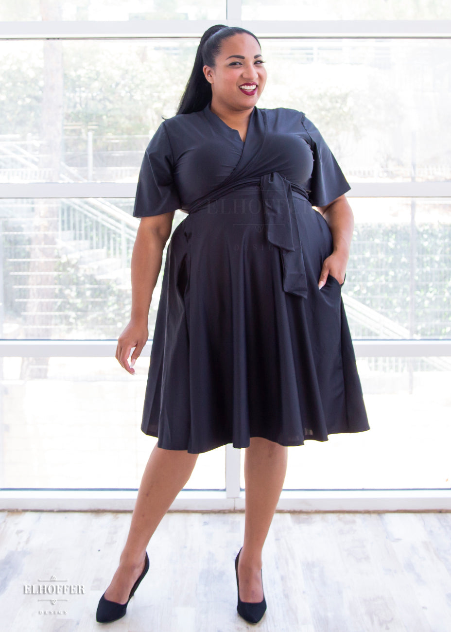 Tas, a medium skinned 2XL model with dark brown hair, is wearing a black knee length wrap dress with flutter sleeves.