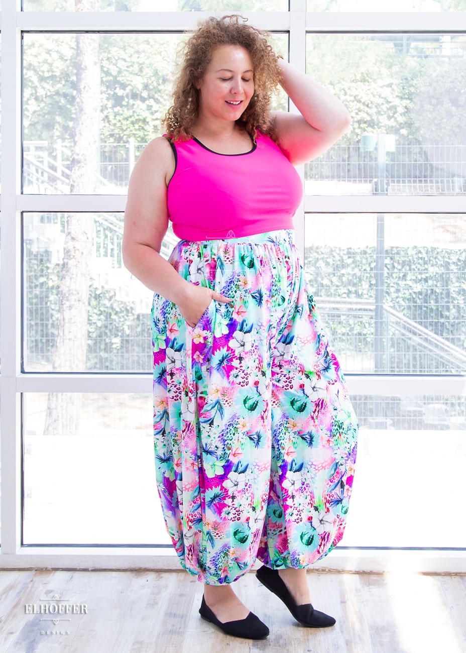 Anastasia, a curly haired, medium-skinned size XL model, is wearing rainbow jungle patterned high waisted full pants gathered at the waist and ankle with a drop crotch seam and side pockets. The pattern is bright blue, pink and green with tropical flowers.