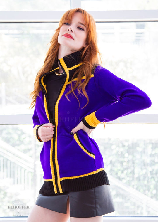 Chloe, a fair skinned XS model with red hair and bangs, is wearing a knit jacket featuring textured sleeves, ribbed kangaroo pockets, a hidden front zipper, and a gorgeous high neckline. The main body of the sweater is bright purple, with black and yellow details.