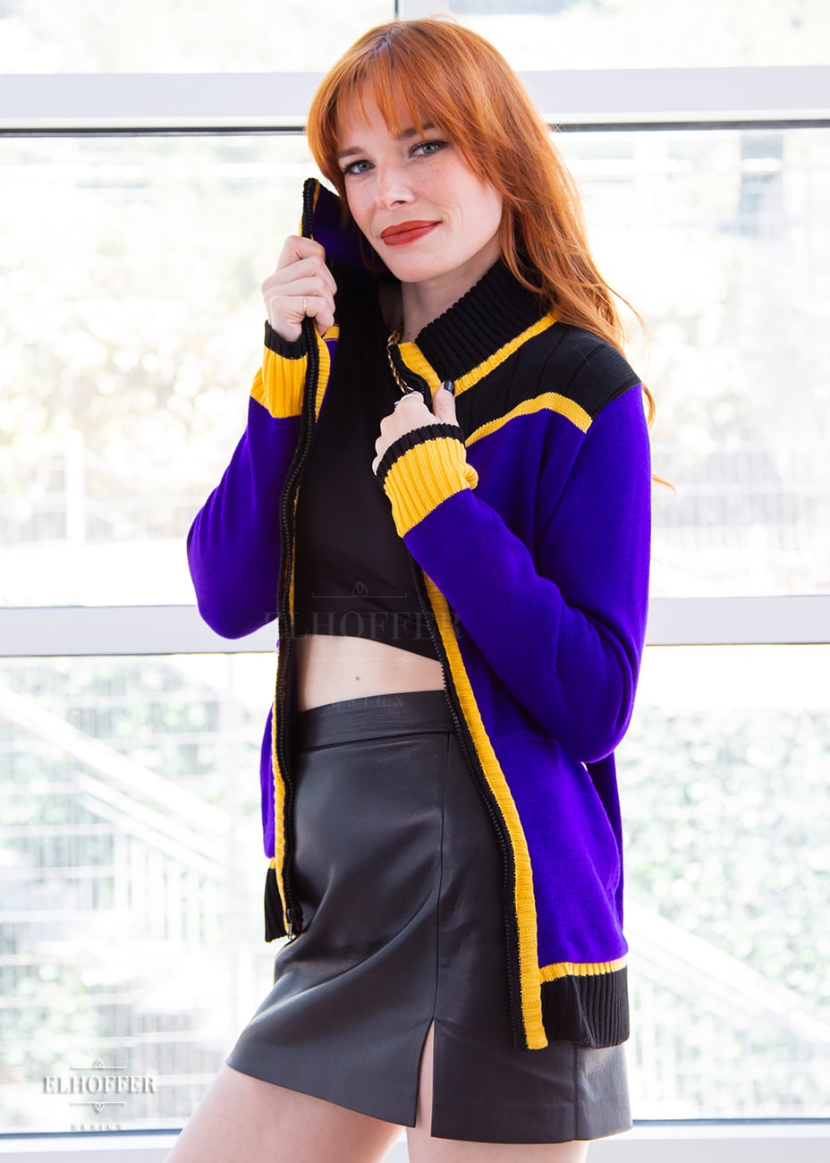 Chloe, a fair skinned XS model with red hair and bangs, is wearing a knit jacket featuring textured sleeves, ribbed kangaroo pockets, a hidden front zipper, and a gorgeous high neckline. The main body of the sweater is bright purple, with black and yellow details.