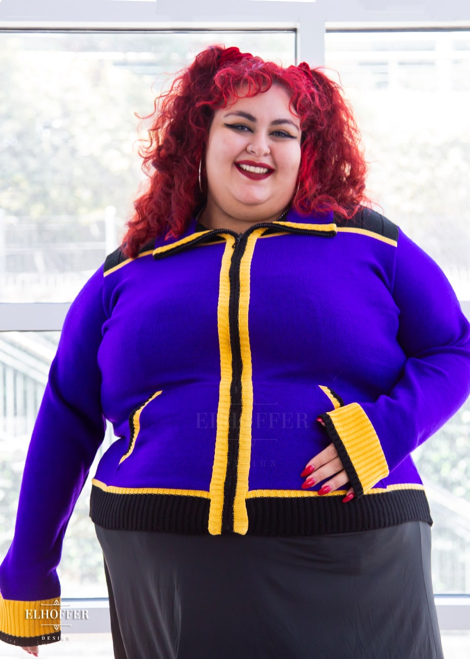Victoria, an olive skinned 4XL model with curly bright red hair, is wearing a knit jacket featuring textured sleeves, ribbed kangaroo pockets, a hidden front zipper, and a gorgeous high neckline. The main body of the sweater is bright purple, with black and yellow details.