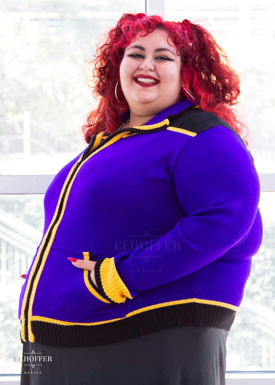 Victoria, an olive skinned 4XL model with curly bright red hair, is wearing a knit jacket featuring textured sleeves, ribbed kangaroo pockets, a hidden front zipper, and a gorgeous high neckline. The main body of the sweater is bright purple, with black and yellow details.