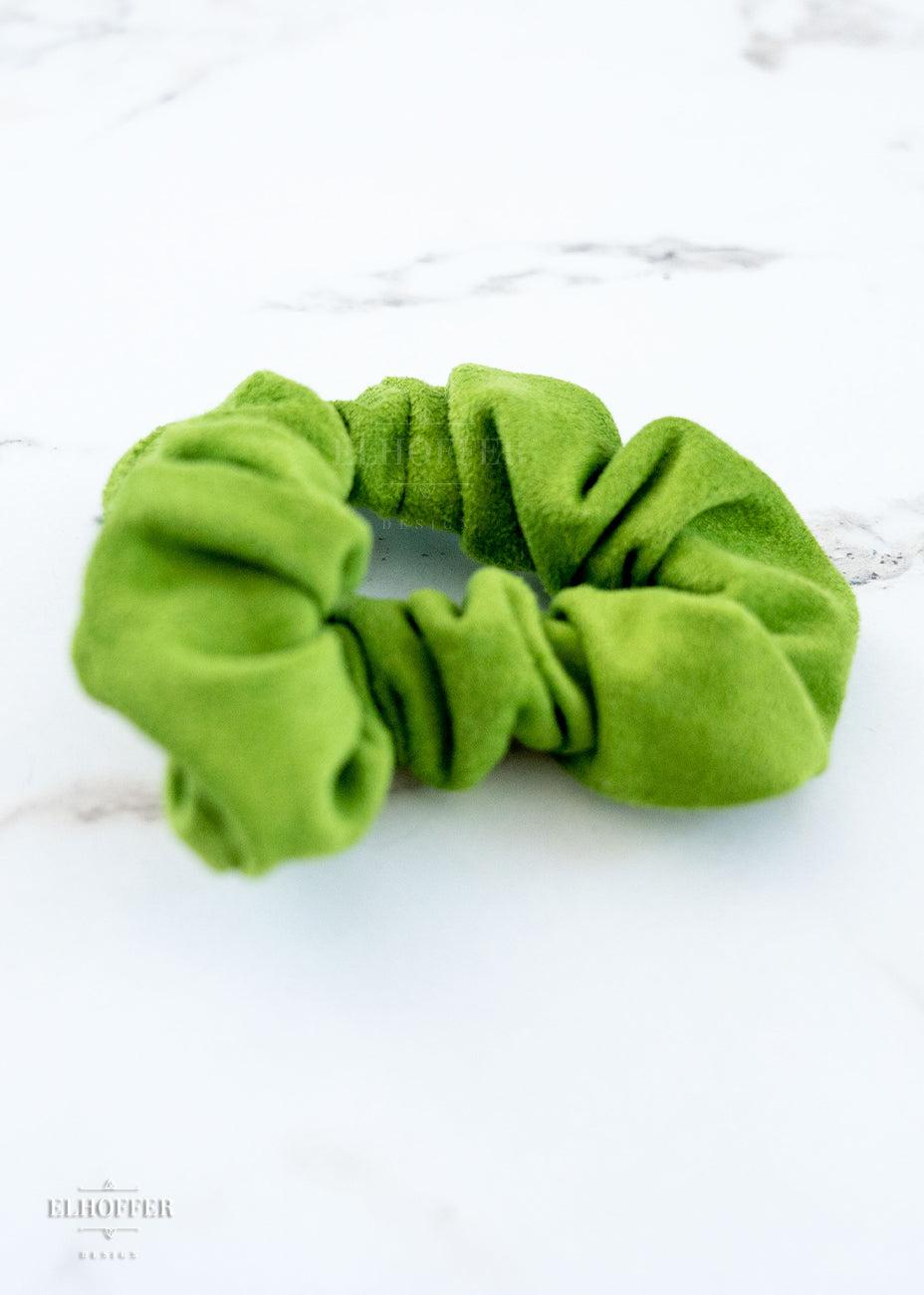 An avocado colored suede scrunchie on a white marbled background.