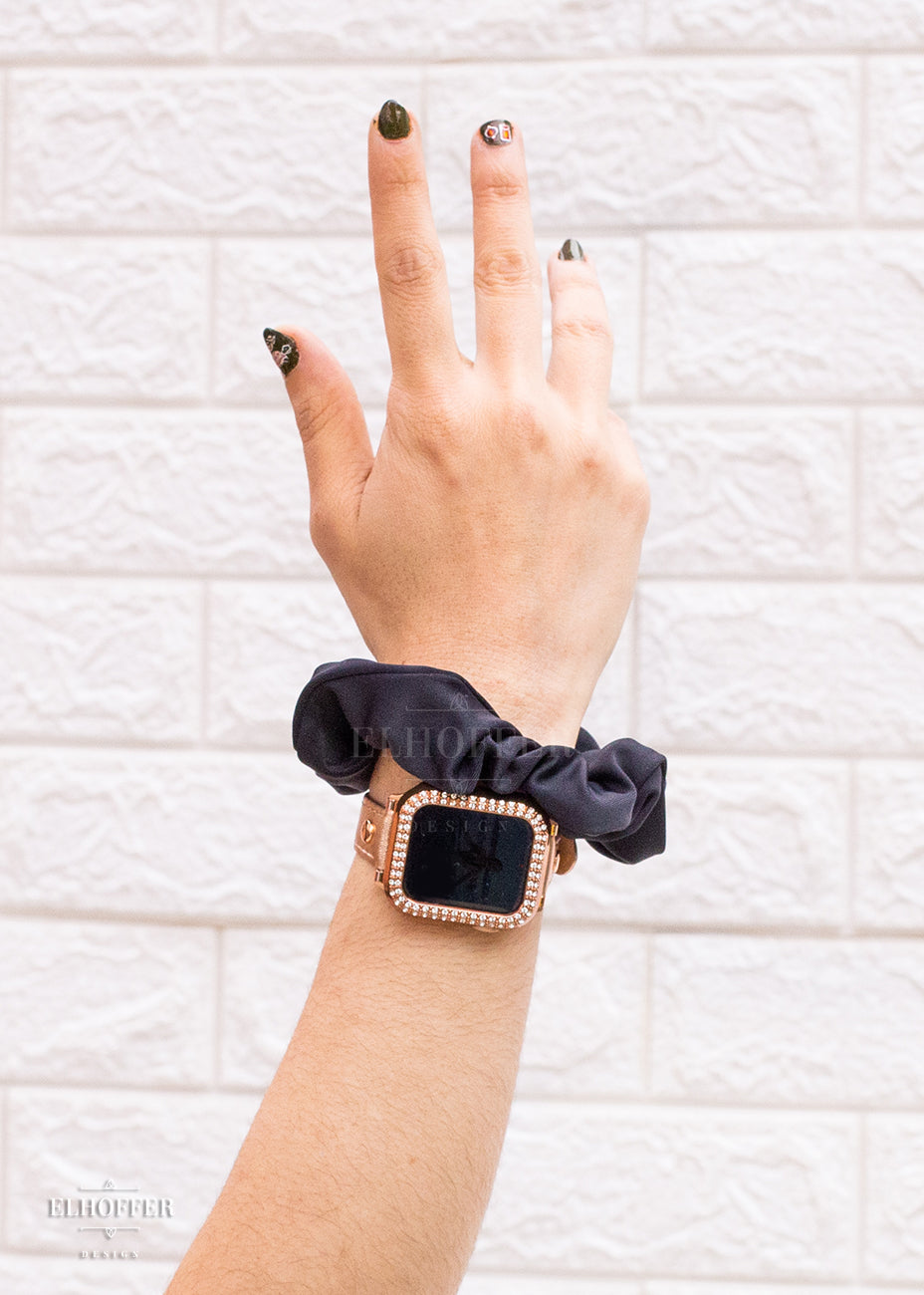 An olive arm with polished nails wearing a solid black scrunchie and rose gold apple watch.