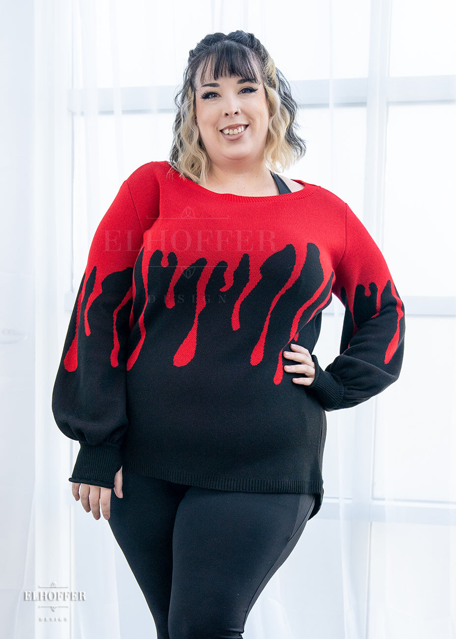 Katie Lynn, a fair skinned 2xl model with short curly black and white hair with bangs, is smiling while wearing an oversize sweater with a bright red blood drip design that looks like it's oozing down onto a black sweater that has long billowing sleeves with thumbholes.