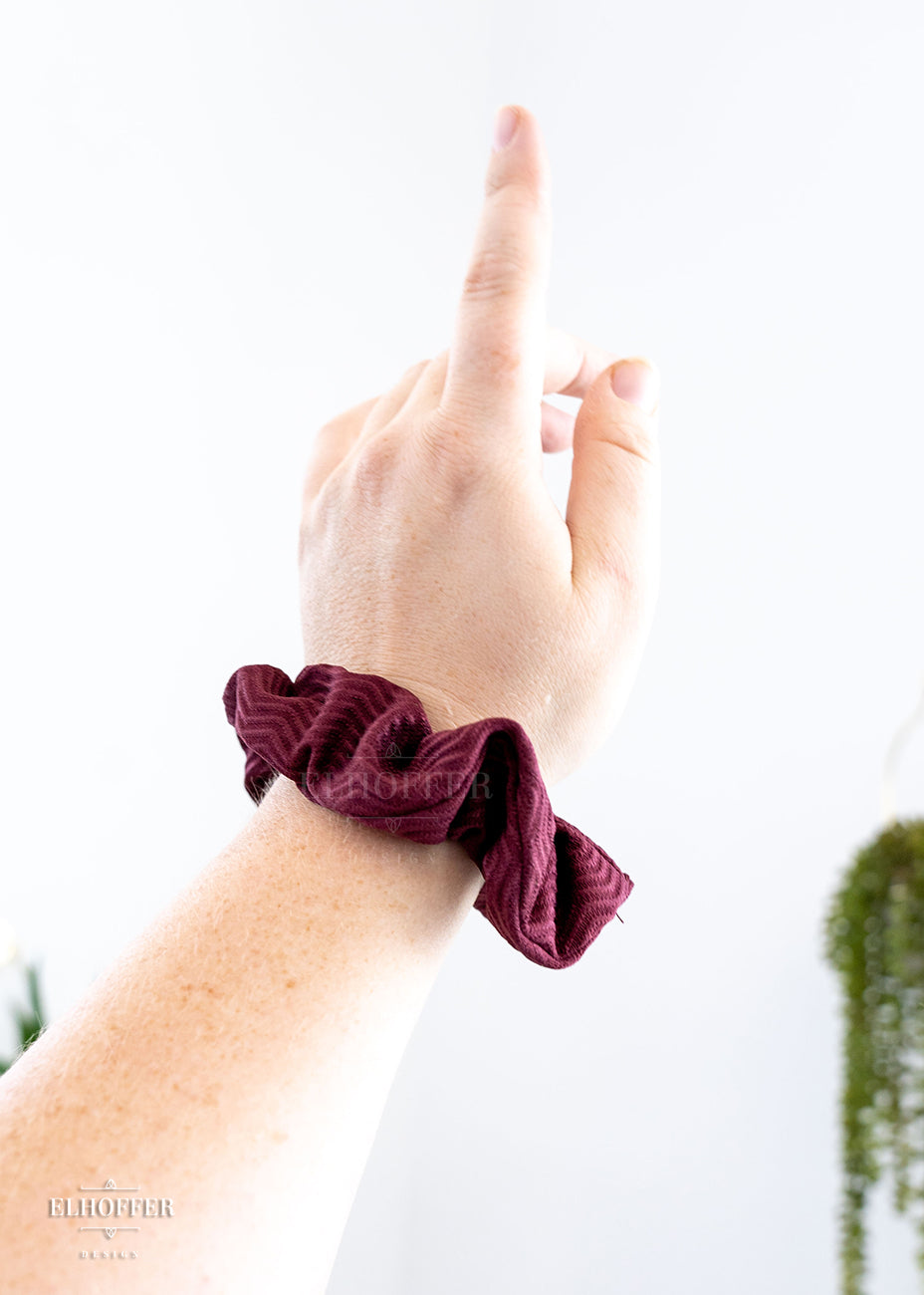 A fair and freckled arm wearing a chaos print scrunchie. The Chaos Red fabric is a narrow chevron textured spandex knit in a dark red shade of burgundy.