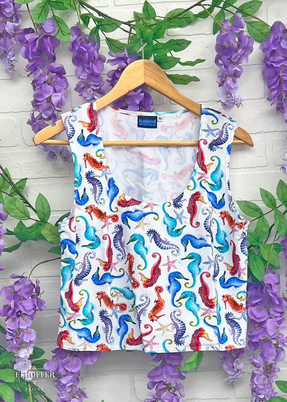 On a hanger in front of wisteria vines, is a sleeveless scoop neck crop top.  The main body of the crop top is white and has rainbow colored seahorses repeating throughout. 