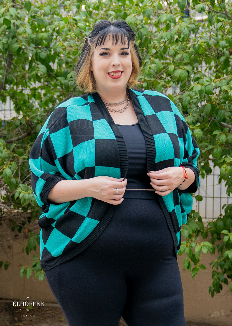 Katie Lynn, a fair skinned 2xl model with short black and blonde hair with bangs, is smiling while wearing a XL-3XL sample of a shrug with a black and green chessboard pattern.  The shrug featured 3/4 sleeves and black ribbing along edges and cuffs.