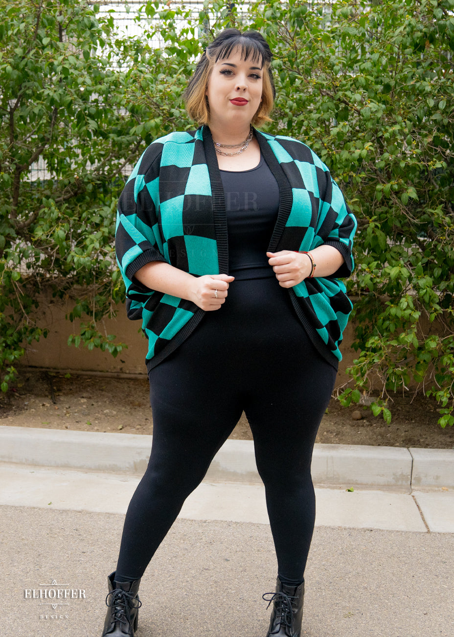 Katie Lynn, a fair skinned 2xl model with short black and blonde hair with bangs, is smiling while wearing a XL-3XL sample of a shrug with a black and green chessboard pattern.  The shrug featured 3/4 sleeves and black ribbing along edges and cuffs.