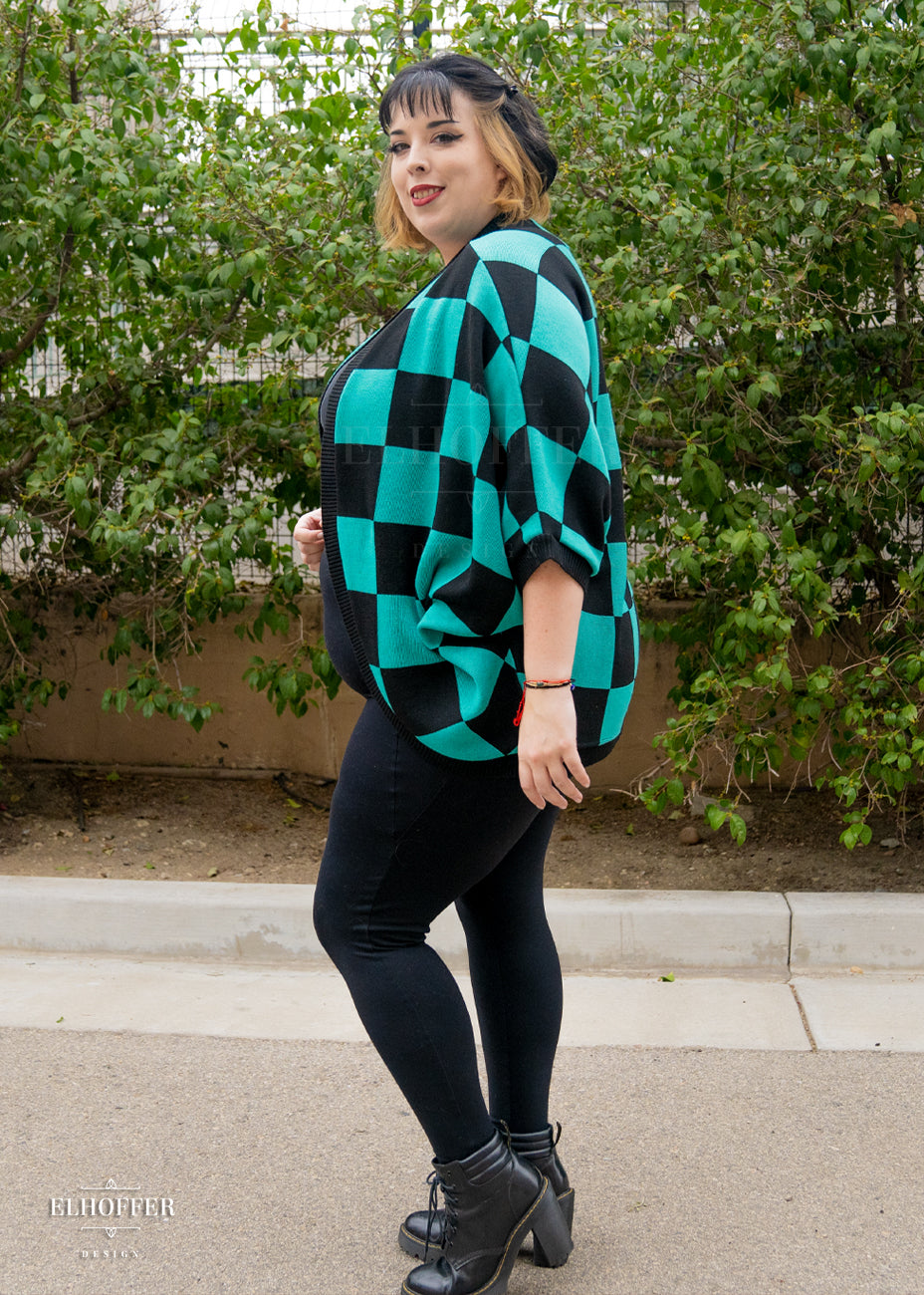 Katie Lynn, a fair skinned 2xl model with short black and blonde hair with bangs, is smiling while wearing a XL-3XL sample of a shrug with a black and green chessboard pattern.  The shrug featured 3/4 sleeves and black ribbing along edges and cuffs.