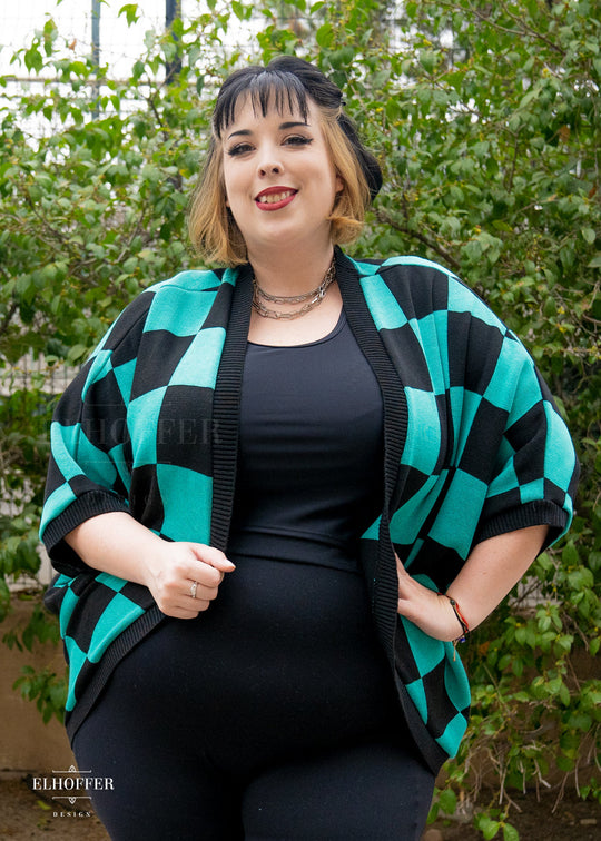 Katie Lynn, a fair skinned 2xl model with short black and blonde hair with bangs, is smiling while wearing a XL-3XL sample of a shrug with a black and green chessboard pattern.  The shrug featured 3/4 sleeves and black ribbing along edges and cuffs.