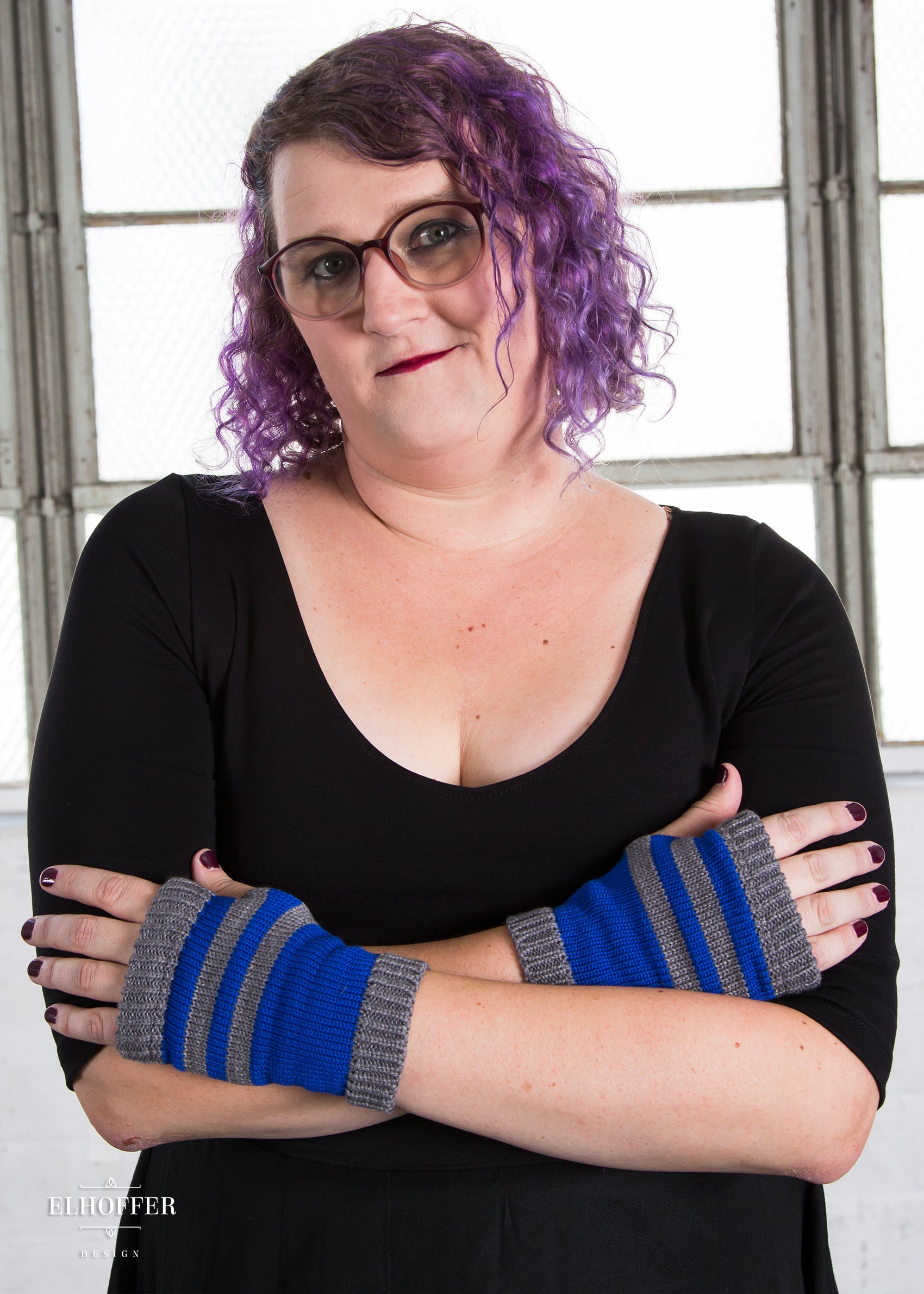 Riley, a size XL model with fair skin and curly purple hair, is wearing a paid of fingerless silver and blue gloves.