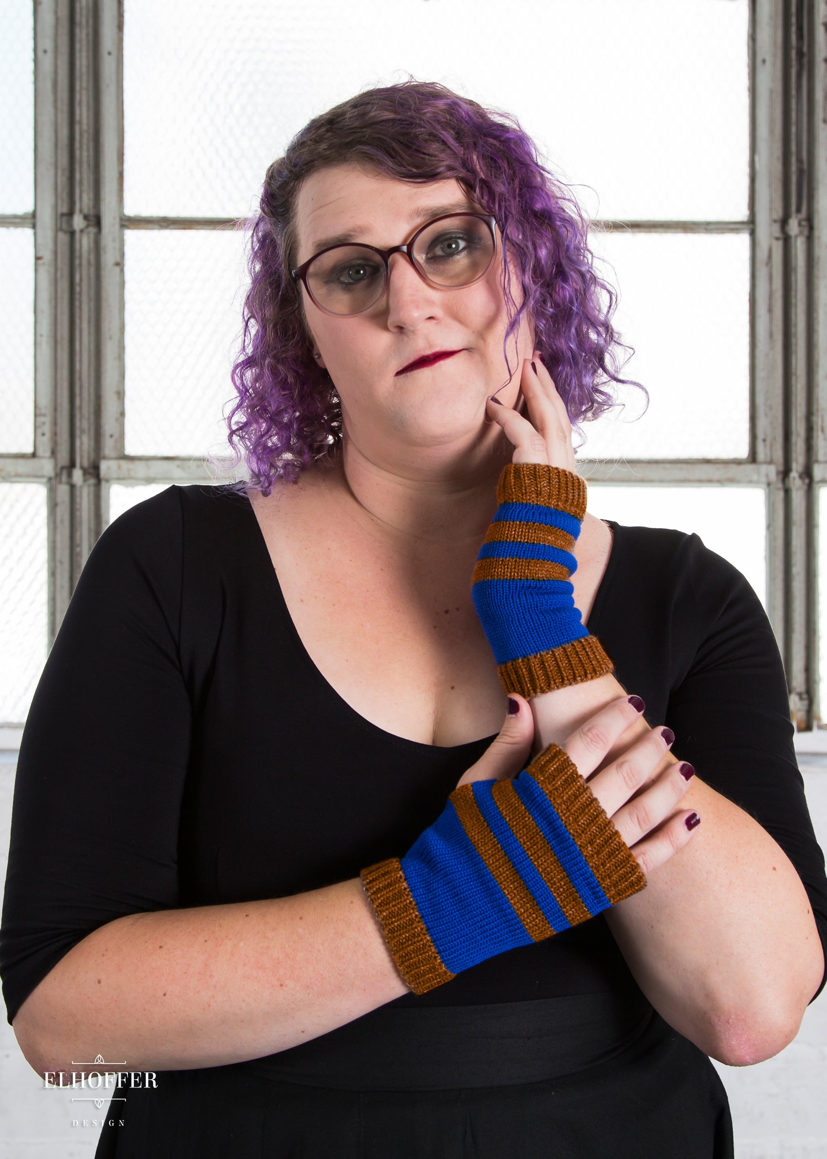 Riley, a fair skinned size XL model with curly purple hair, is wearing a pair of bronze and blue striped fingerless gloves.